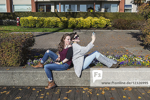 Zwei junge Frauen sitzen Rücken an Rücken und benutzen ihre Smartphones und machen Blödsinn; New Westminster  British Columbia  Kanada