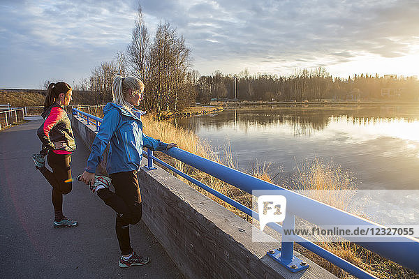 Two young women stretching on a trail at the water's edge; Anchorage  Alaska  United States of America