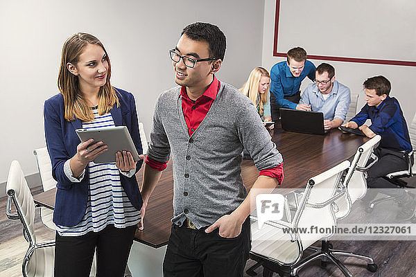 Zwei junge Geschäftsleute schauen auf ein Tablet  während sie in einem Konferenzraum mit Gleichaltrigen zusammenarbeiten; Sherwood Park  Alberta  Kanada