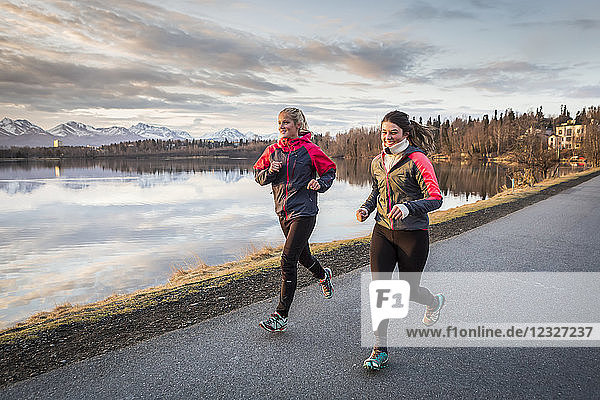 Two young women running on a trail at the water's edge with mountains in the distance; Anchorage  Alaska  United States of America