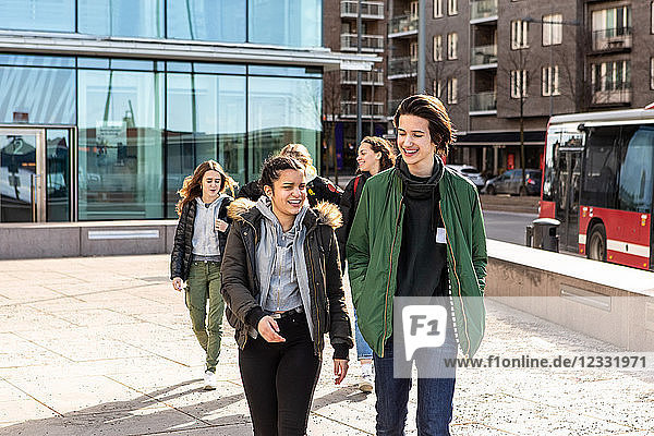 Smiling multi-ethnic teenagers wearing warm clothing walking on footpath in city during sunny day