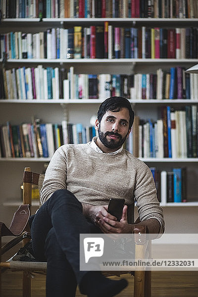 Portrait of young man holding smart phone while sitting against bookshelf