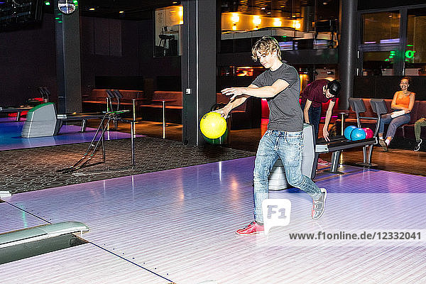Full length of teenage boy holding ball on parquet floor against friends at bowling alley