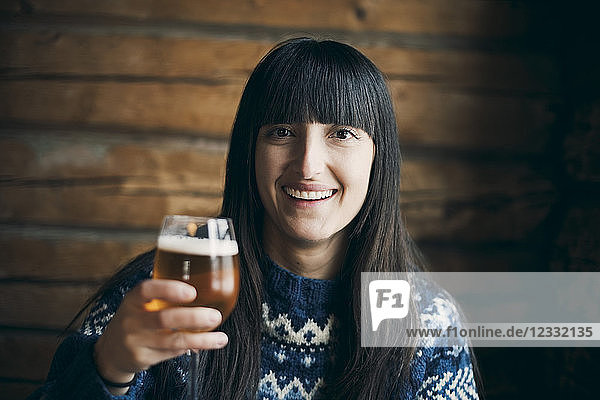 Portrait of smiling woman holding drinking beer glass while sitting at log cabin