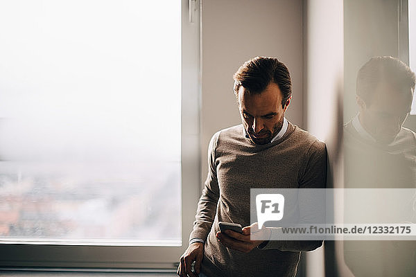 Businessman leaning on wall while using smart phone against window at creative office