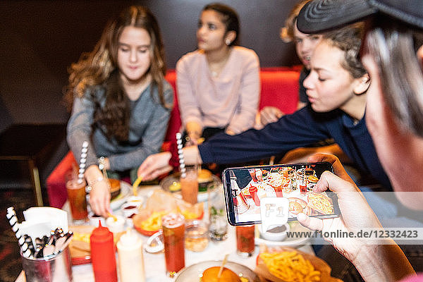 Cropped image of teenage boy sitting with friends while photographing food and drinks on table at restaurant