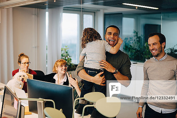 Cheerful businessman carrying son while standing amidst colleagues at creative office