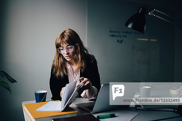 Confident young businesswoman reading document while sitting with laptop at creative office