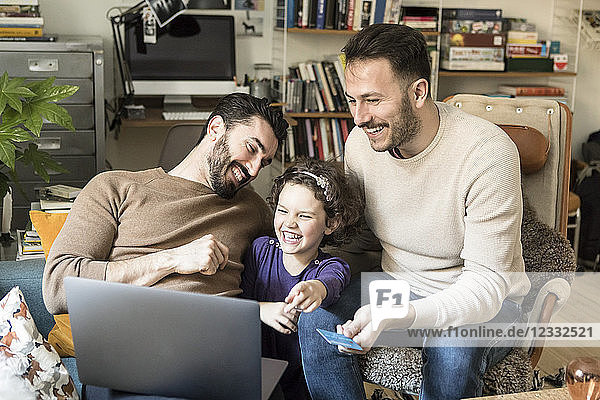 Happy fathers with daughter shopping online on laptop through credit card while sitting on chairs at home