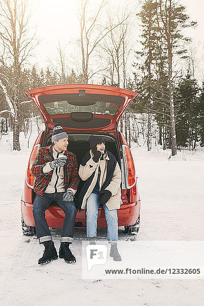 Full length of friends having coffee while sitting in car trunk on snowy field