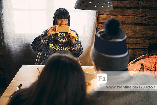 Woman photographing friends through mobile phone while sitting in log cabin