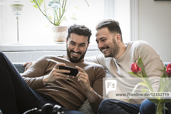 Smiling young men looking at smart phone while sitting on sofa at home