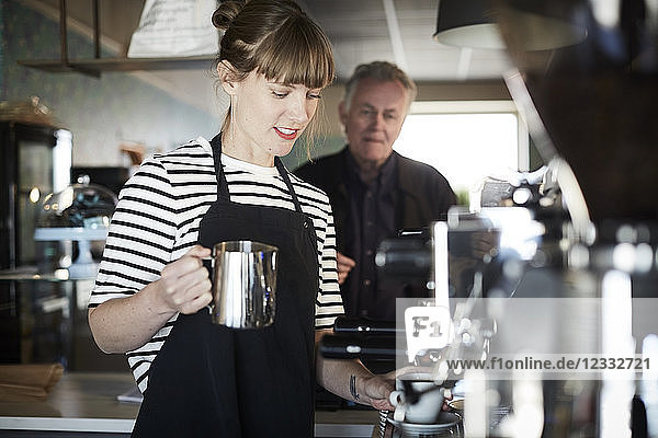 Female barista holding pitcher while making coffee with customer standing in background at cafe