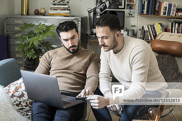 Gay couple shopping online with credit card through laptop while relaxing on chairs at home