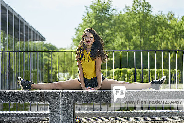 Portrait of smiling young woman doing splits on bench outdoors