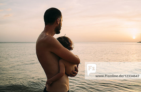 Father holding little daughter on his arms while watching sunset together at the sea