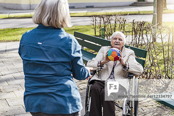 Woman taking care of old woman in wheelchair playing with ball in park