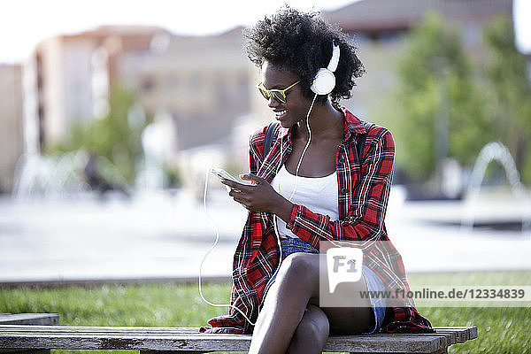 Smiling young woman sitting on bench in city park listening music with headphones