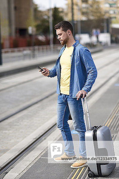Young man waiting at a station with smartphone in his hand and trolley