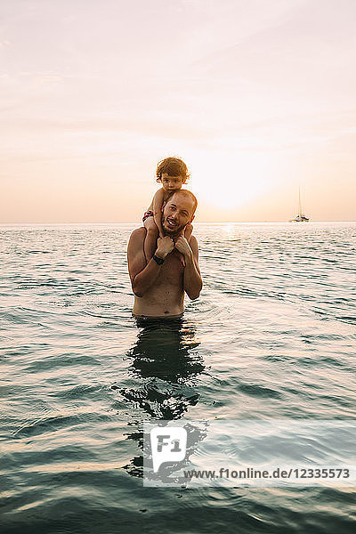Father carrying his little daughter on shoulders in the sea by sunset