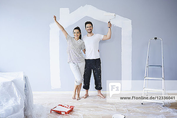 Portrait of cheerful couple painting in new apartment with house shape on wall