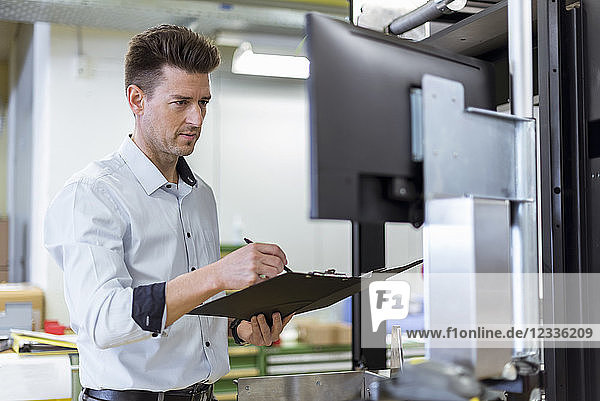 Man with clipboard in factory looking at screen