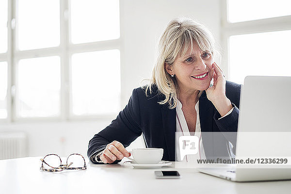 Smiling mature businesswoman with cup of coffee working on laptop at desk in the office