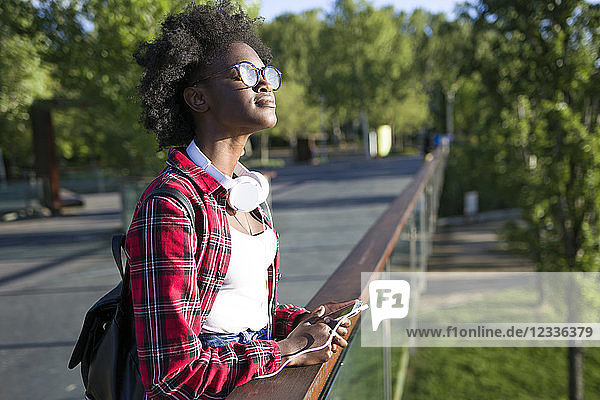 Young woman with headphones and cell phone standing on a bridge enjoying sunlight