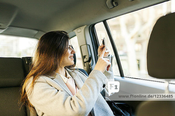 Smiling young woman sitting on backseat of a car taking picture with cell phone