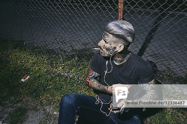 Tattooed young man with earbuds and smartphone smoking a cigarette at wire mesh fence