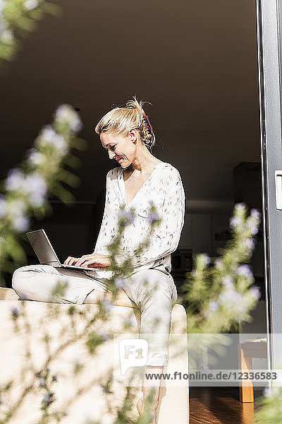 Smiling mature woman sitting at open terrace door using laptop