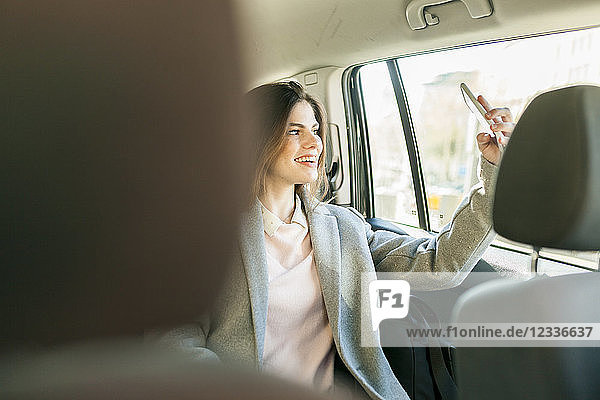 Smiling young businesswoman sitting on backseat of a car taking selfie with cell phone