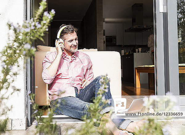 Smiling mature man sitting at open terrace door listening music with headphones