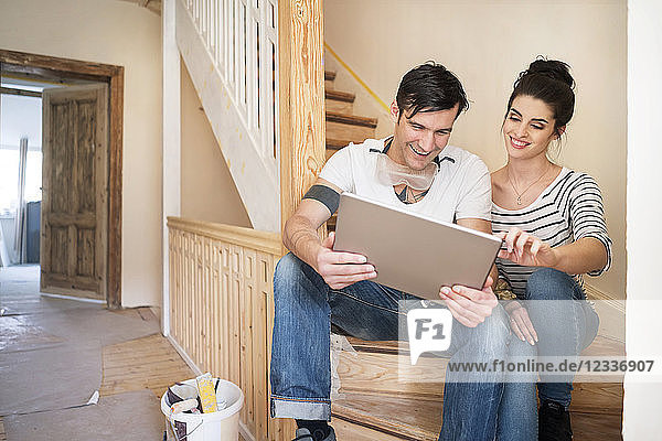 Young couple refurbishing new home  sitting on stairs using digital tablet