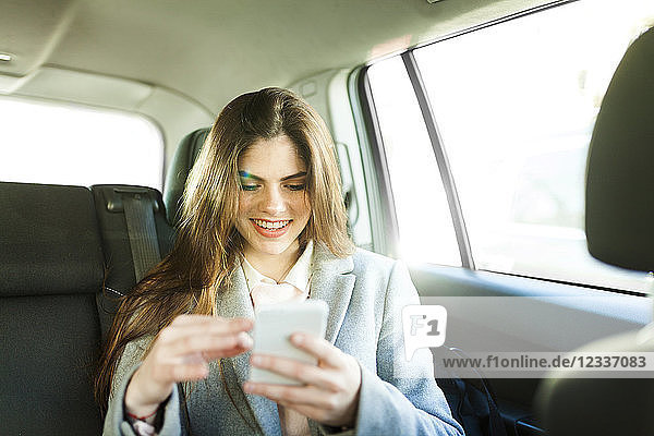 Portrait of smiling young businesswoman sitting on backseat of a car looking at cell phone
