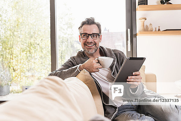 Portrait of smiling man with cup of coffee sitting on couch with tablet