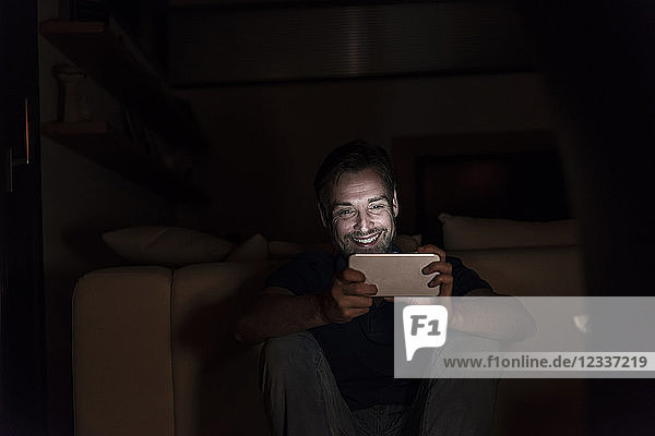 Laughing man sitting in the dark at home looking at smartphone