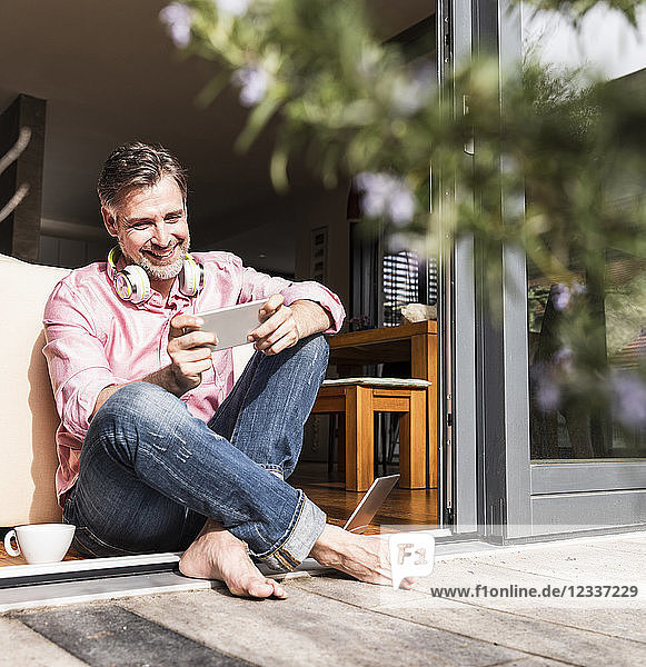 Smiling mature man sitting at open terrace door looking at smartphone
