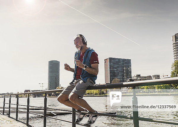 Happy athletic mature man with headphones sitting at the riverside in the city