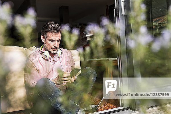 Mature man sitting at open terrace door using smartphone