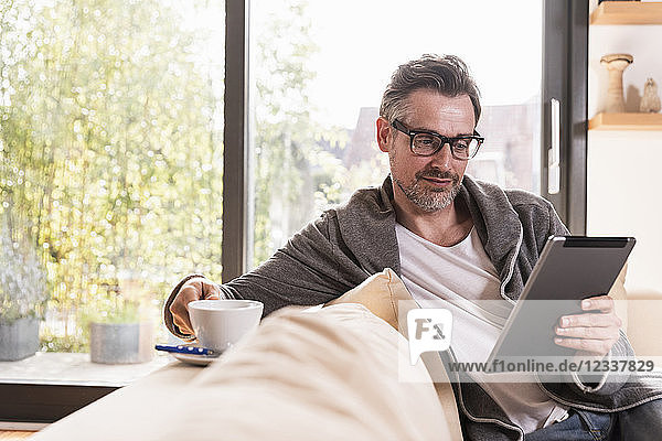 Portrait of mature man with cup of coffee sitting on couch using tablet