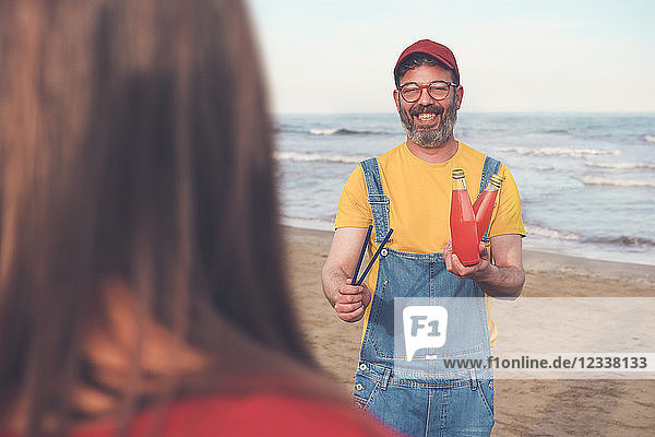 Happy man in dungarees on the beach offering soft drink to woman