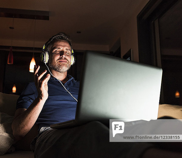 Man sitting on couch at home listening music with headphones and laptop