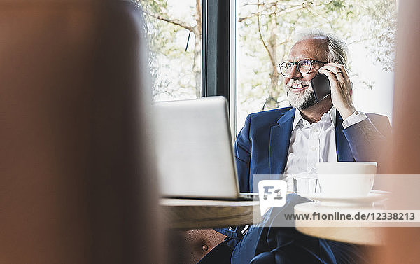 Mature businessman sitting at table in a cafe using cell phone and laptop
