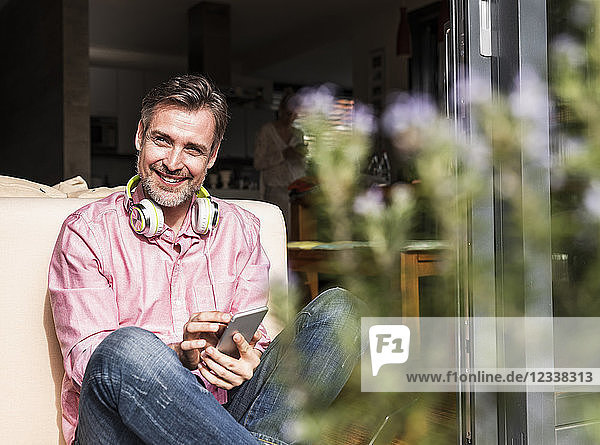 Portrait of smiling mature man sitting at open terrace door using smartphone