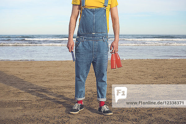 Man in dungarees standing on the beach holding bottles of soft drinks