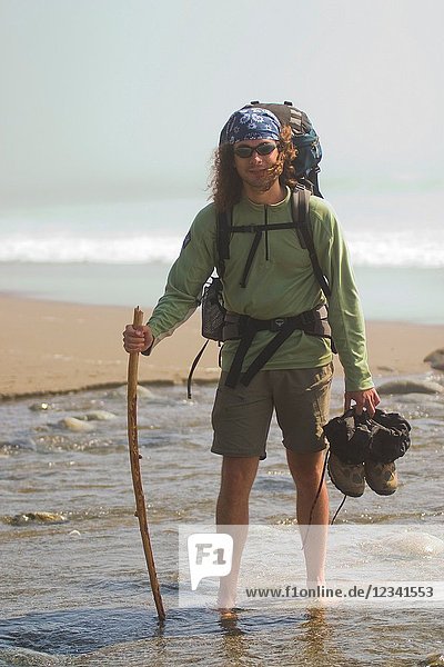 Backpacker on the beach of the West Coast Trail with a stick and boots in his hands standing in the low water. West Coast Trail near Pacific Rim National Park near Carmanagh Lighthouse British Columbia Canada