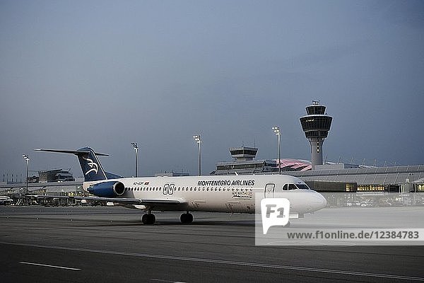 Montenegro Airlines  Fokker  F100  rollt vor dem Terminal 1 mit Tower  in der Abenddämmerung  Flughafen München  Oberbayern  Bayern  Deutschland  Europa