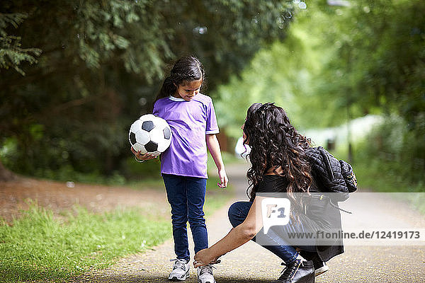 Mother tying shoelace of daughter holding soccer ball