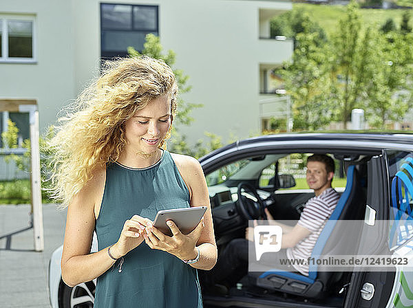 Smiling young woman using tablet with man sitting in electric car in background